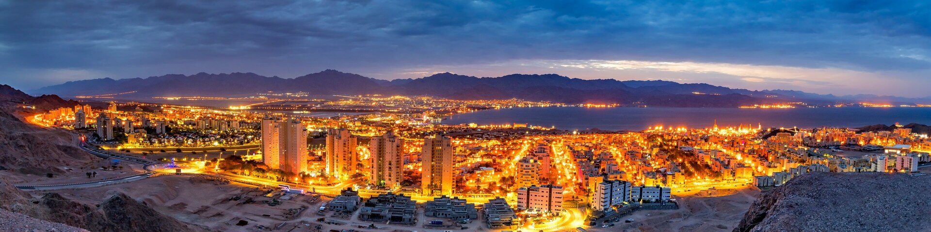 A cityscape at dusk with illuminated buildings and a mountainous backdrop.