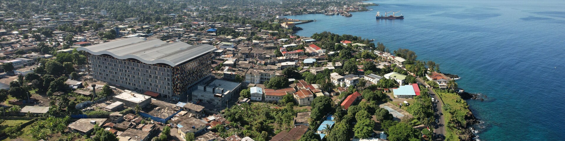 An aerial view of a coastal town with a large industrial building, dense housing, and a harbor with ships.