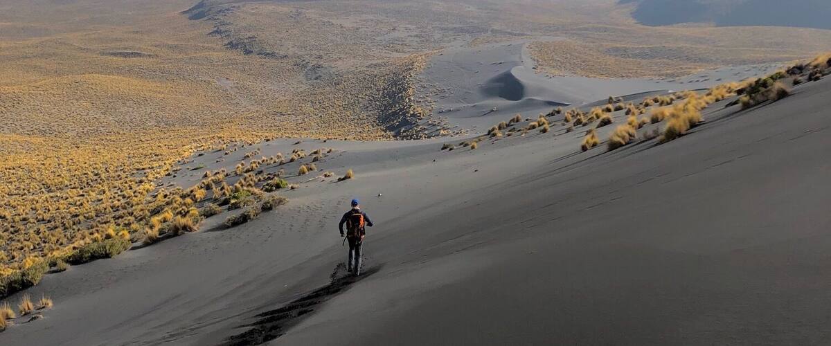 A person hiking on a desert trail with sparse vegetation and a mountain in the background.