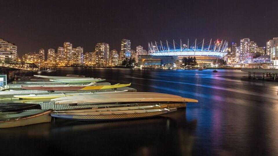 A waterfront with boats docked, a city skyline, and a stadium with lights.