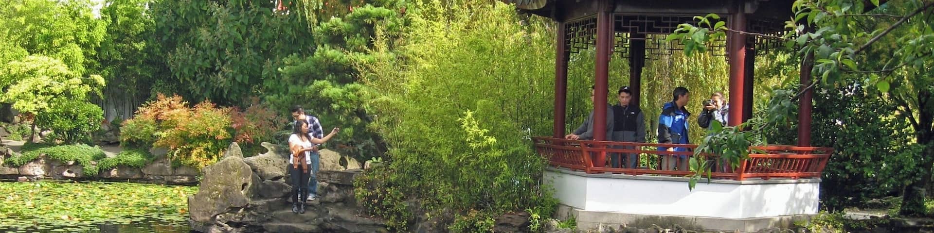 A traditional Chinese pavilion by a pond with lush greenery and a stone bridge.
