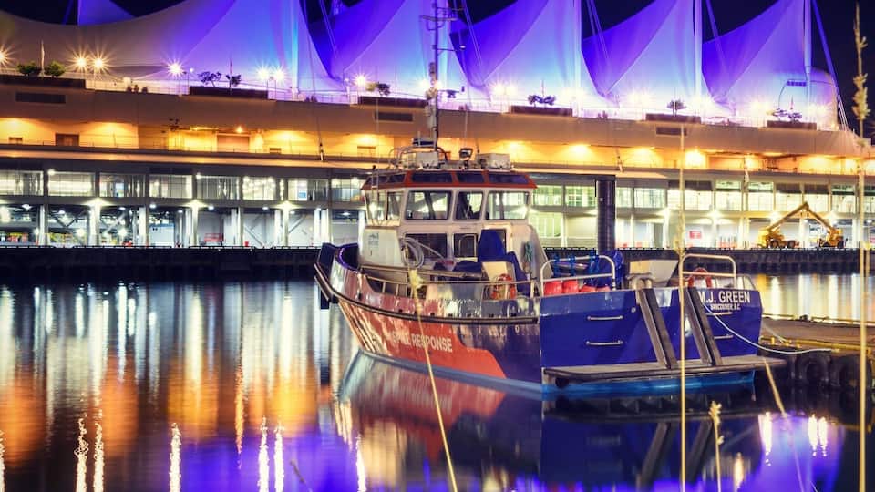 A marina at night with boats docked, a building with a distinctive roof, and a calm water reflecting the lights.