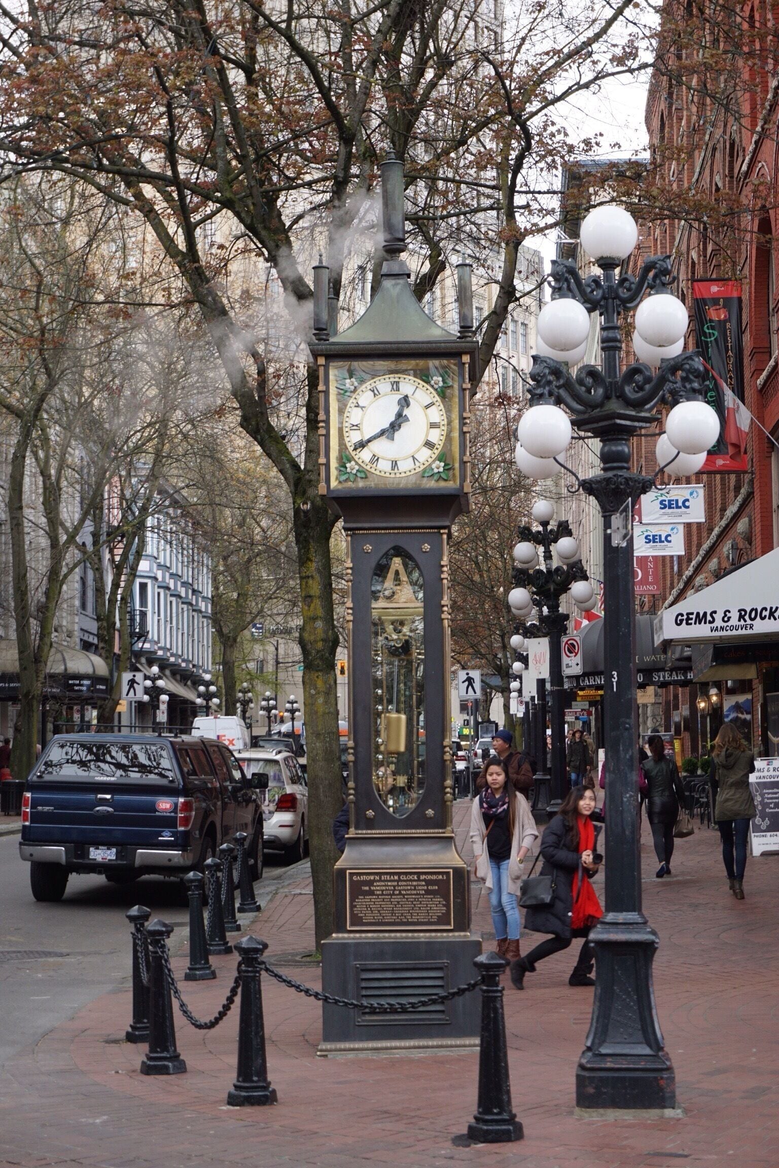 Uma rua com uma torre do relógio, uma estátua e um edifício com a placa 'GEMS & ROCKS'.