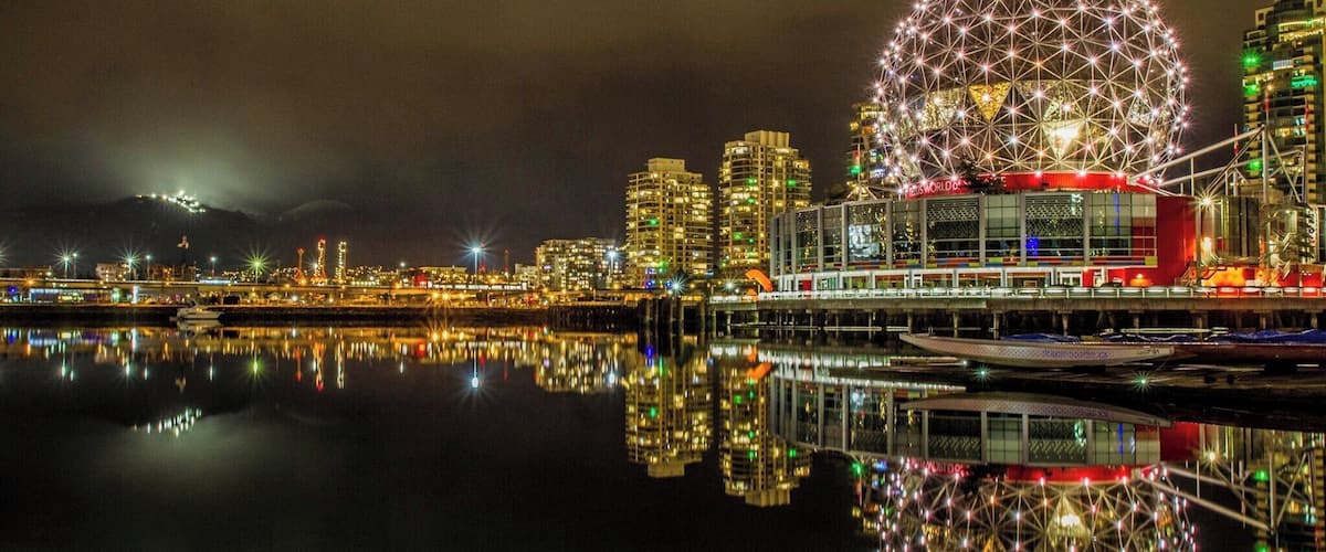 A cityscape at night with a large Ferris wheel reflecting on the water, illuminated buildings, and a bridge.