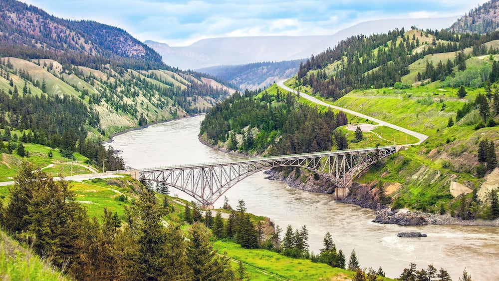 A steel bridge over a river, surrounded by green hills and trees.
