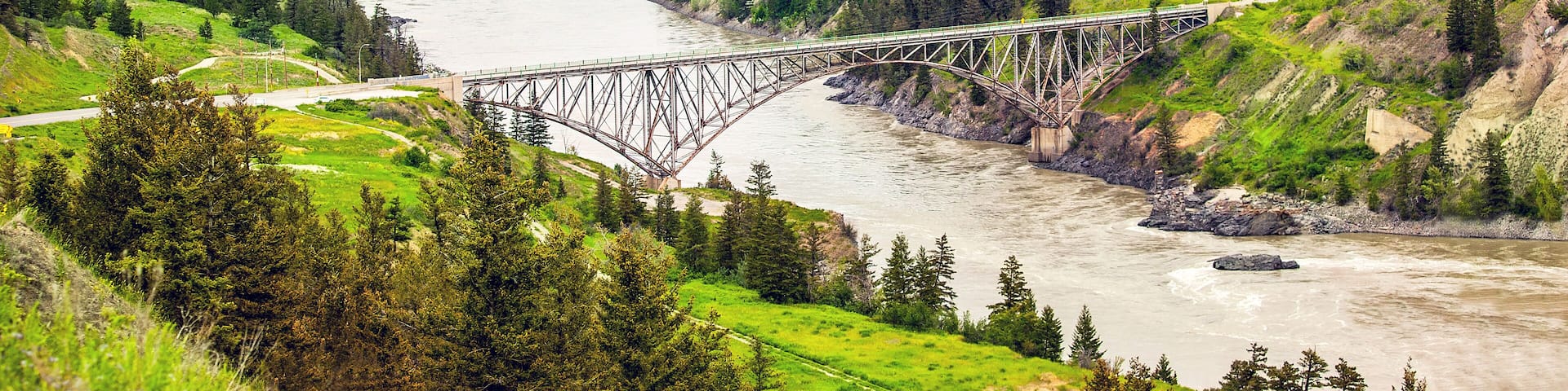 A steel bridge over a river, surrounded by green hills and trees.