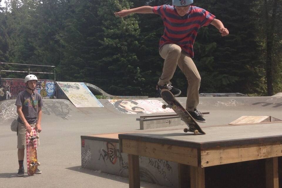 A skateboarder performing a trick at a skatepark with graffiti on the walls and trees in the background.