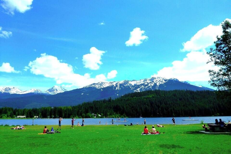 Un lago con gente descansando sobre la hierba, una cordillera al fondo y un cielo azul despejado.