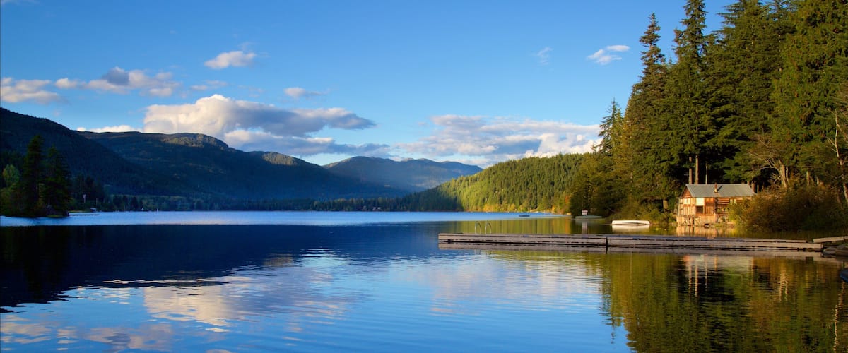 A calm lake with a wooden dock, surrounded by dense forests and mountains under a clear sky.