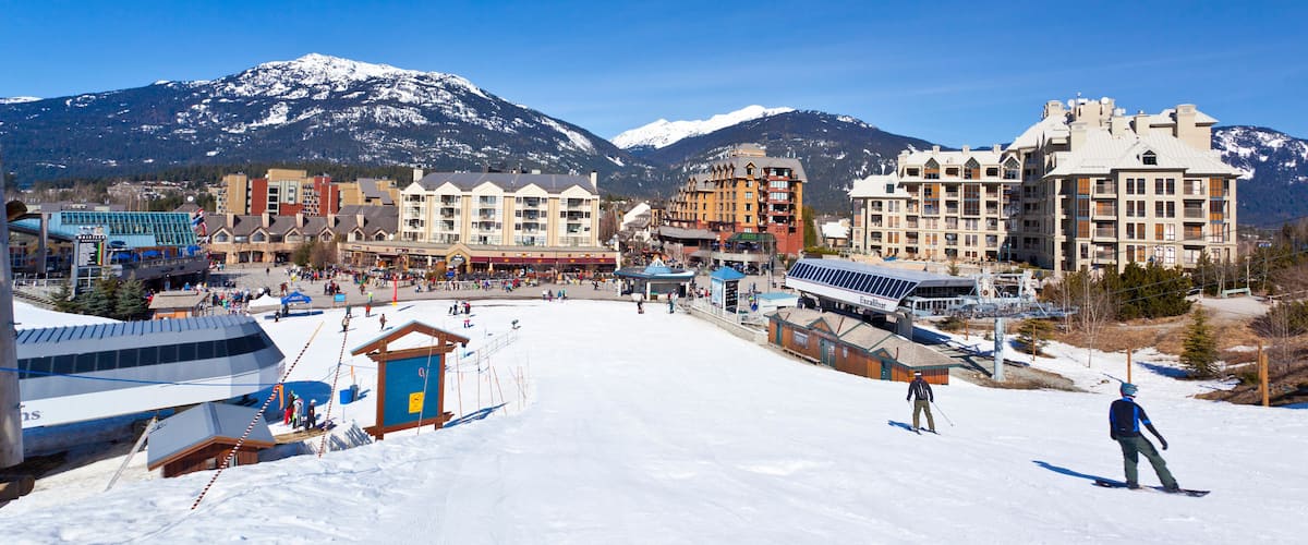A snowy ski resort with buildings, a ski lift, and mountains in the background.