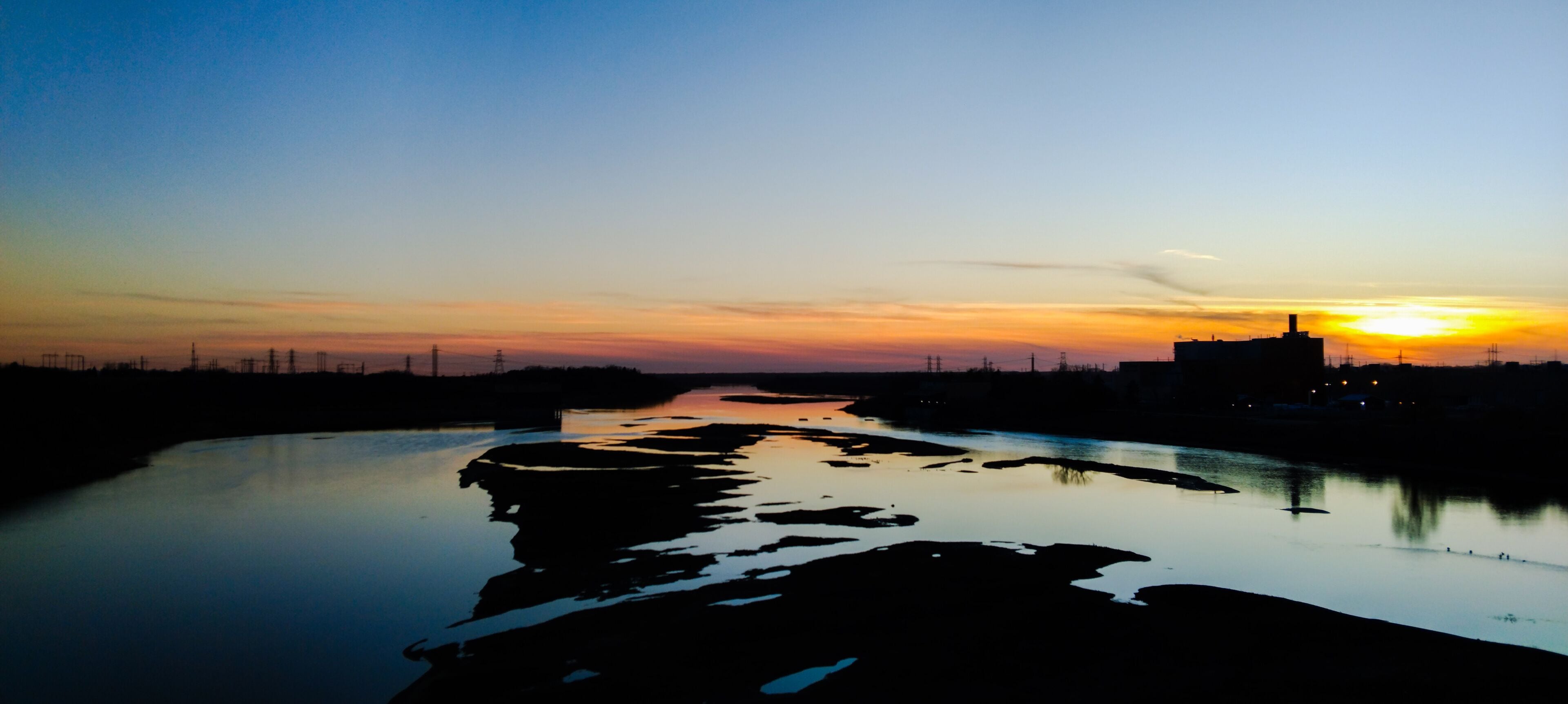 Um rio refletindo o pôr do sol, silhueta distante de uma cidade e linhas de energia elétrica na margem.