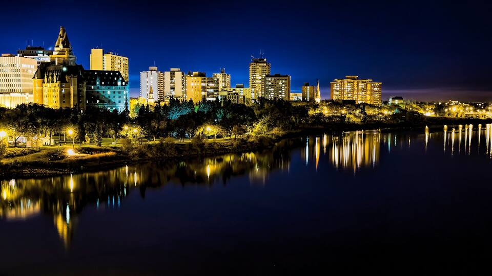 A nighttime cityscape with illuminated buildings and a bridge over a calm river reflecting the lights.