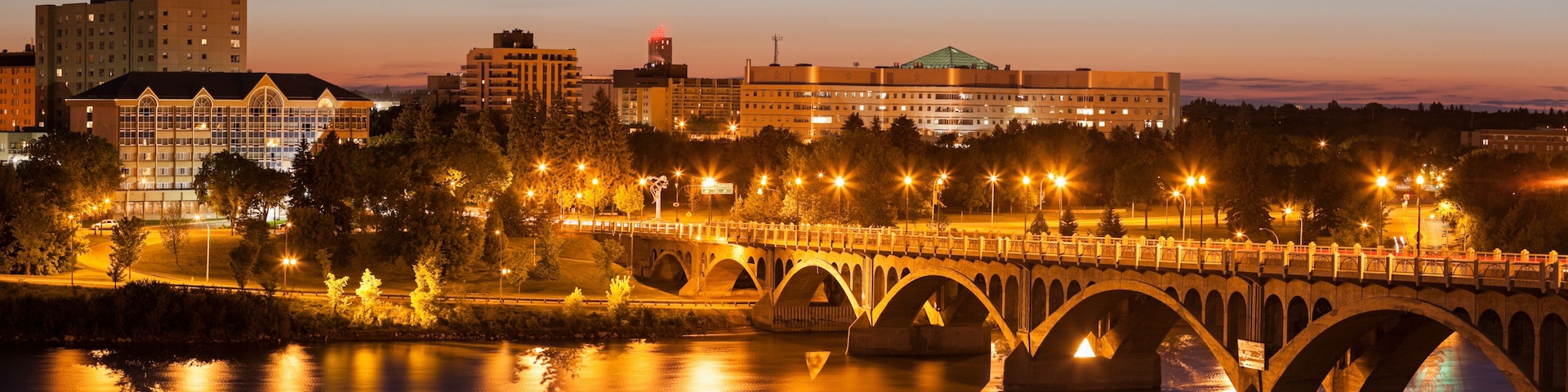 A bridge over a river at dusk with city buildings in the background.