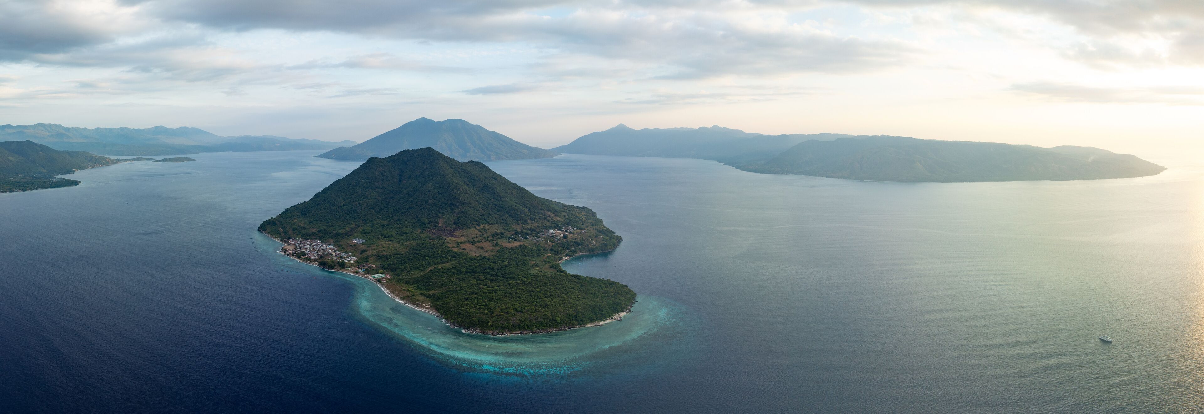 An aerial view of a tropical island with a central peak, surrounded by clear blue waters and distant mountains.