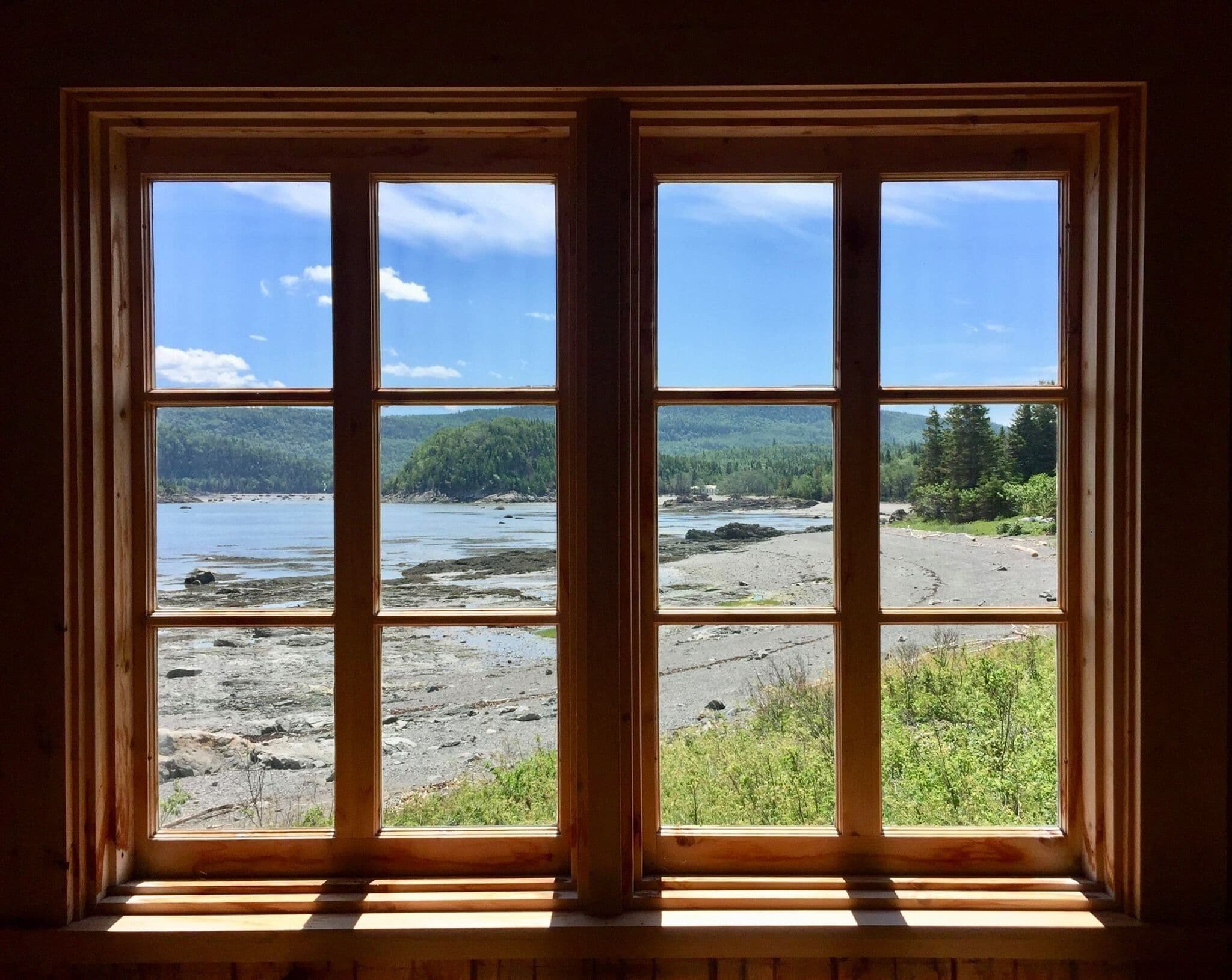 A wooden window with a view of a rocky shoreline and forested hills.