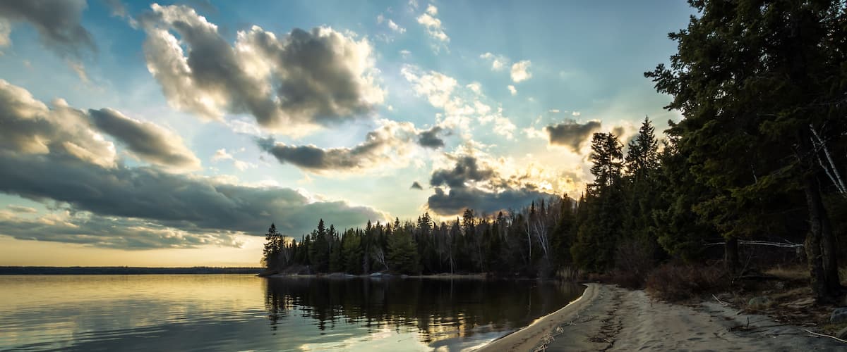 A tranquil lakeside with a sandy beach, dense forest, and a cloudy sky.