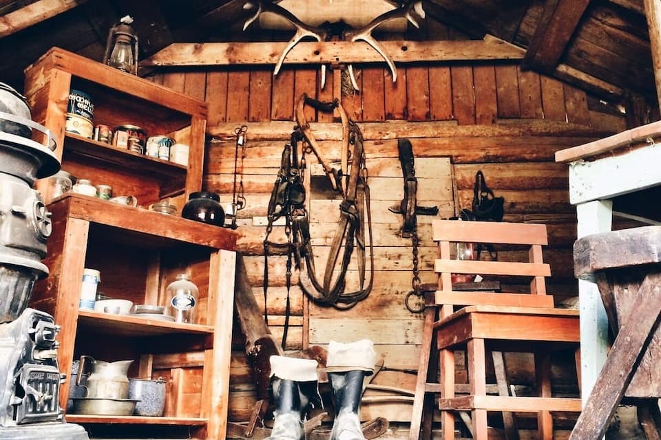A rustic wooden interior with shelves holding jars and tools, a pair of boots, and antlers on the ceiling.