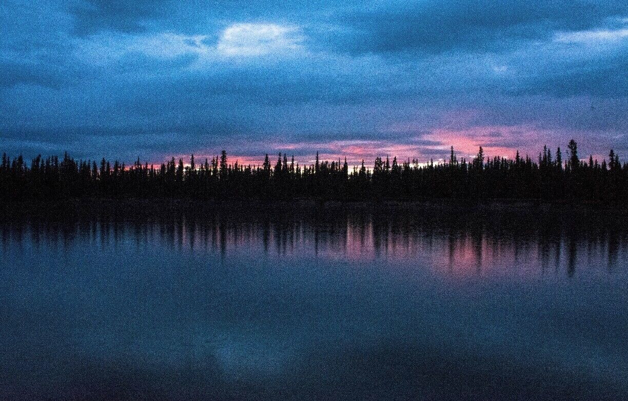 A calm lake reflecting a forest at dusk with a sky transitioning from blue to pink hues.