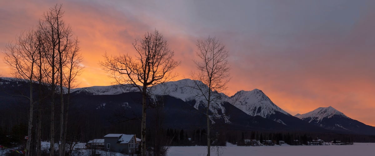 A snow-covered mountain range at sunset with bare trees in the foreground.