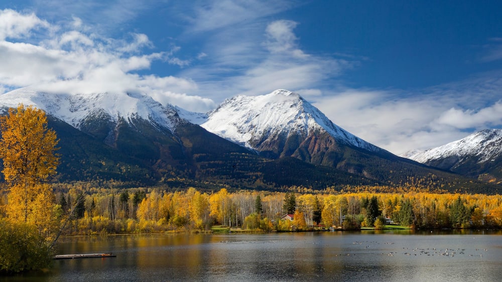 Um lago rodeado por árvores em tons de outono e montanhas cobertas de neve.