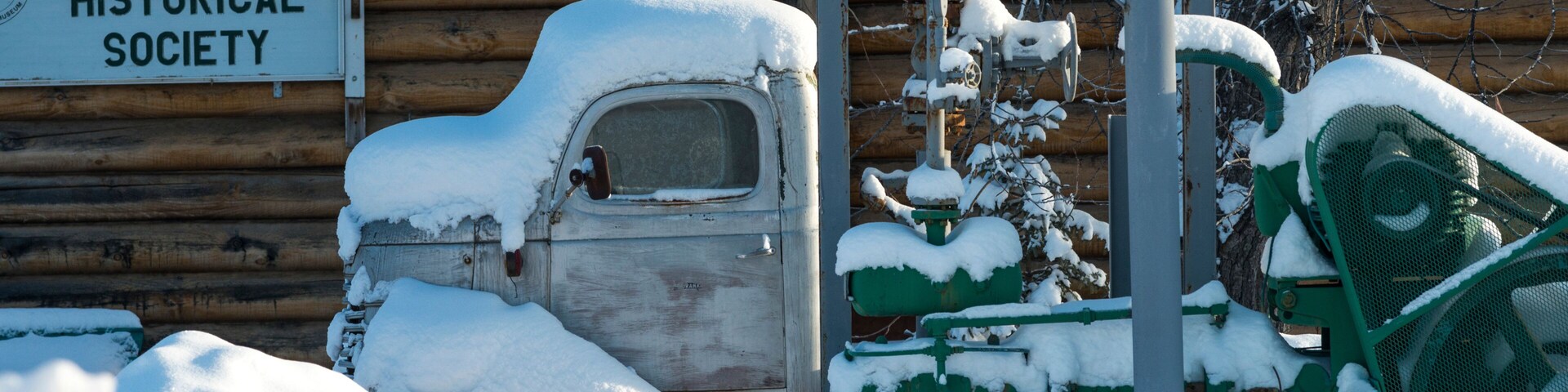Un camión y equipo cubiertos de nieve frente a un edificio de madera con un cartel del Fort Nelson Historical Society.