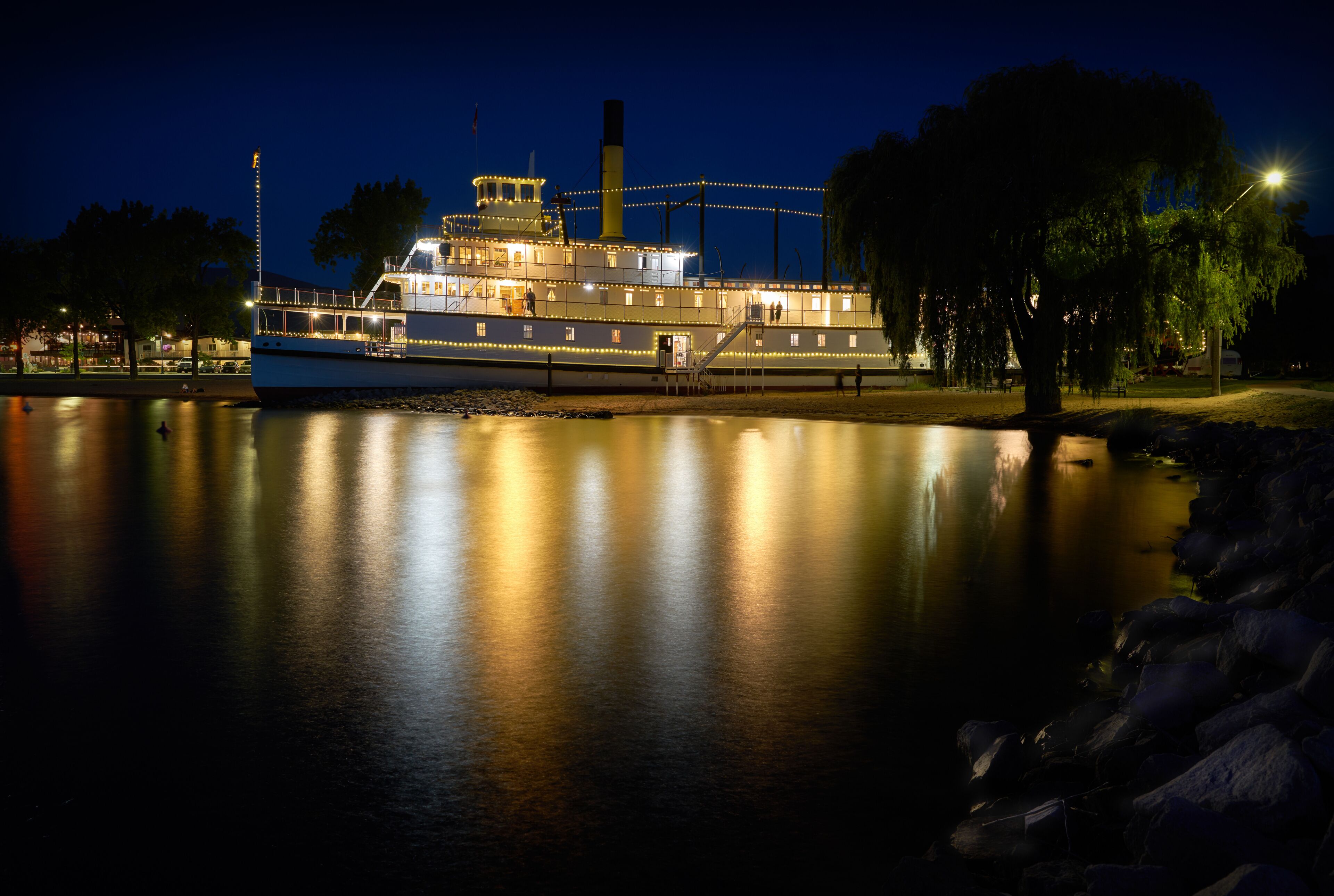 A riverboat with lights reflecting on the water, surrounded by trees and a dock.