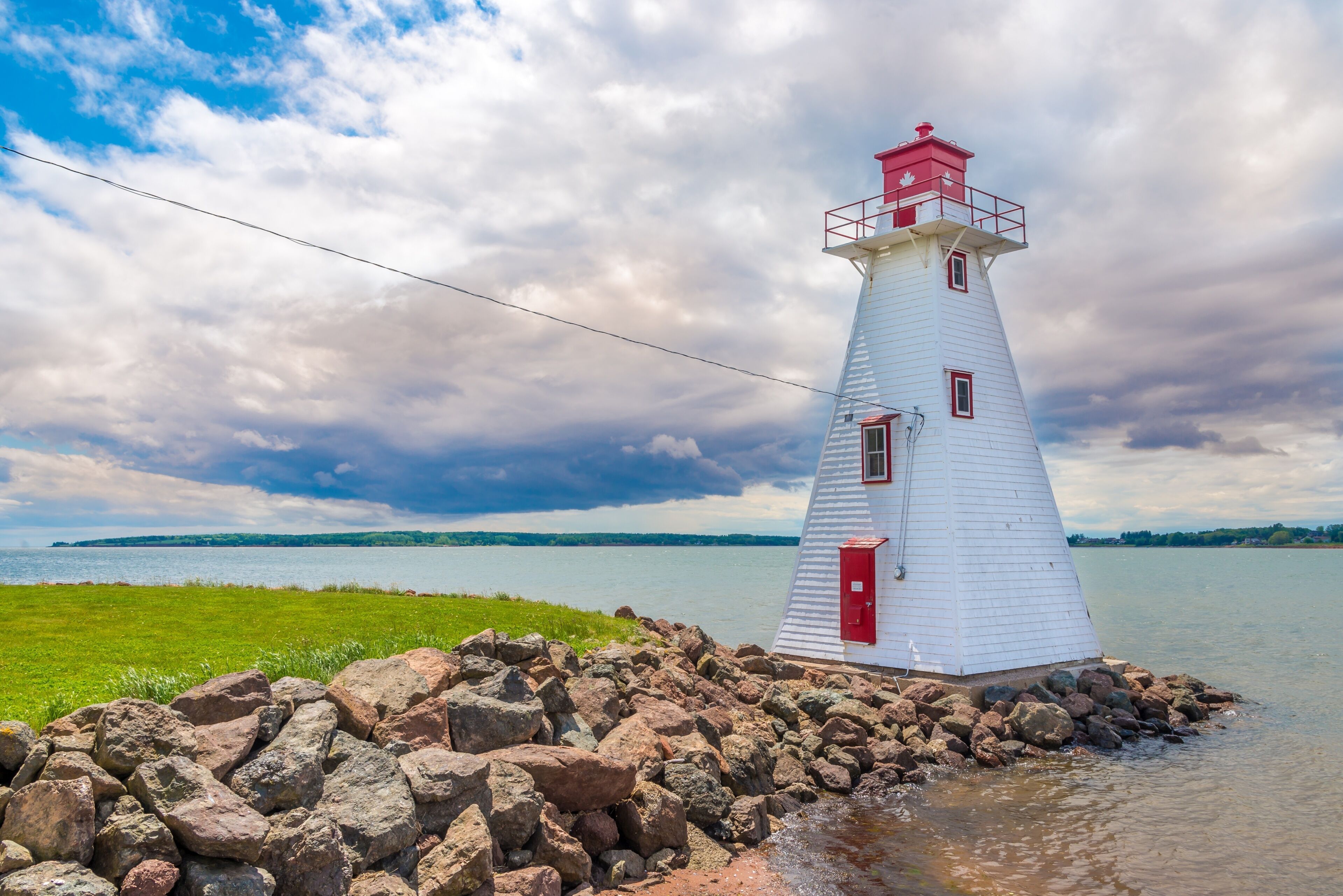A white lighthouse with red accents, situated on a rocky shoreline with a grassy area in the foreground and a body of water in the background.