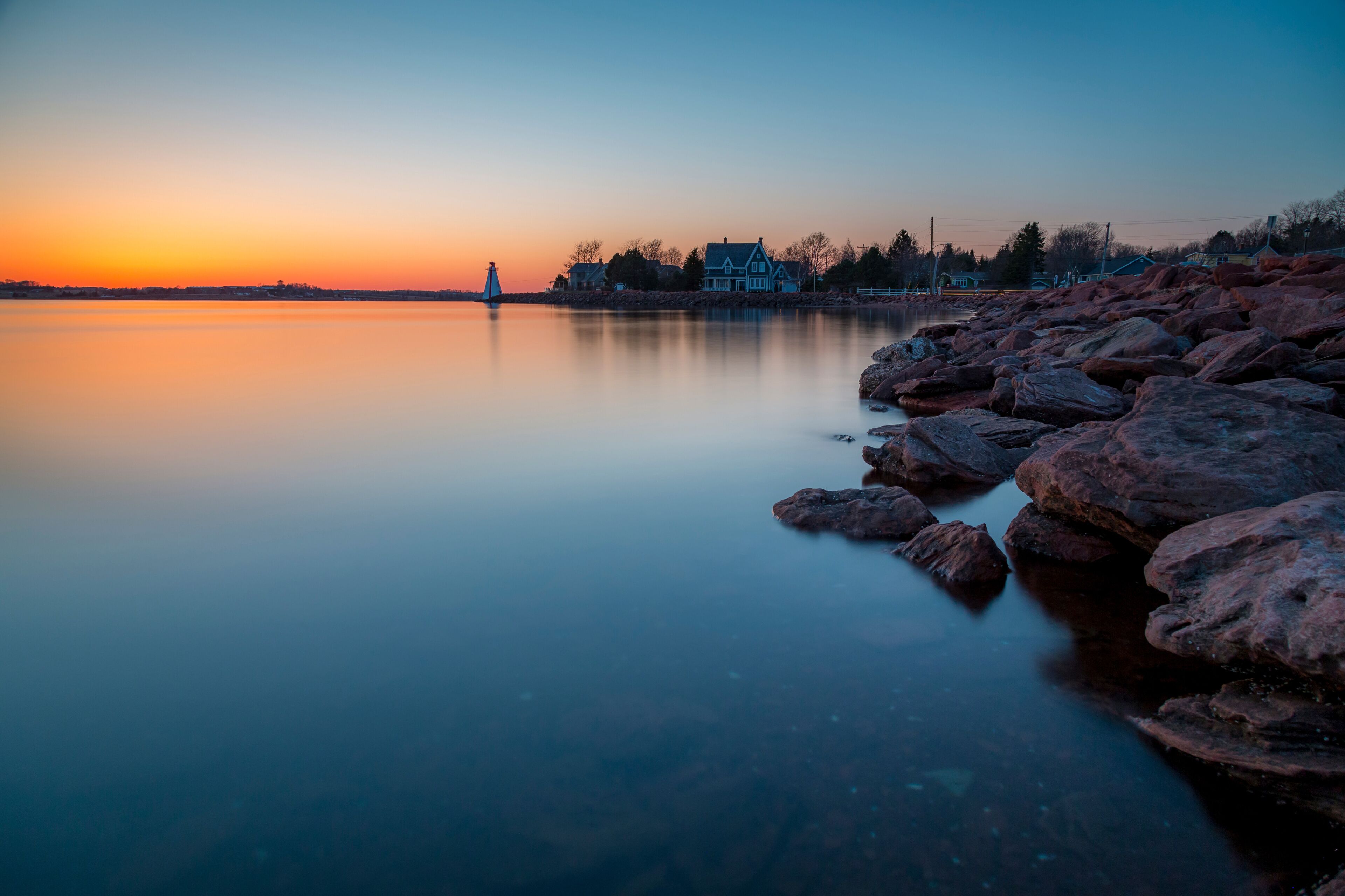 A calm lake at sunset with a lighthouse, rocky shore, and a house in the background.