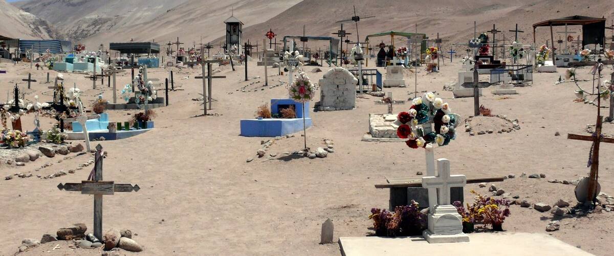 A cemetery with numerous graves, crosses, and headstones, set against a backdrop of mountains.