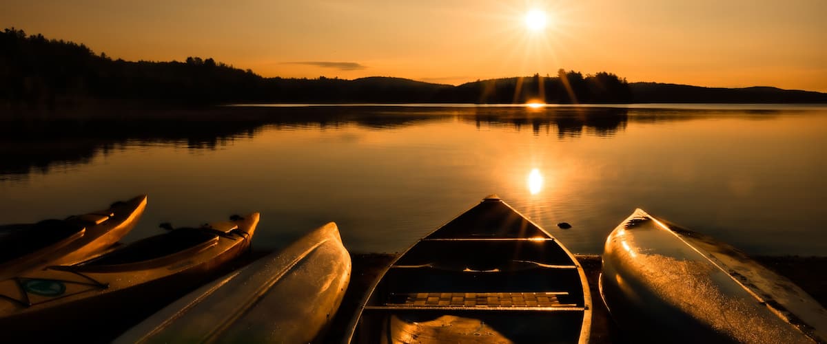 A calm lake at sunrise with kayaks lined up on the shore and a forested hill in the background.