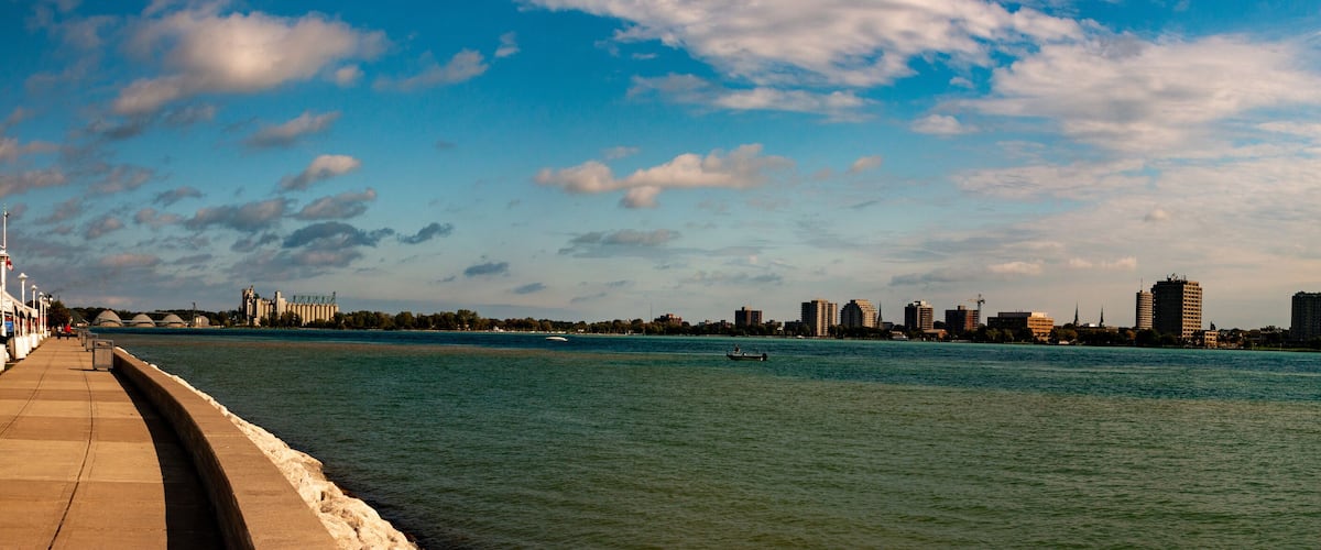 A river with a concrete walkway on the left, a boat in the water, and a city skyline with various buildings in the background.