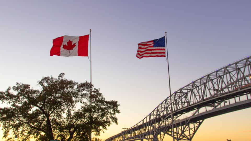 Two flags, a Canadian and an American, flying on flagpoles with a bridge in the background.