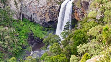 Uma cascata desce por um penhasco rochoso, cercada por vegetação verde exuberante.