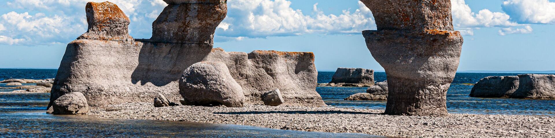 Breathtaking limestone sculptures rise from a shallow coastal pool at Mingan Archipelago National Park Reserve, framed by a clear blue sky adorned with dramatic clouds.
