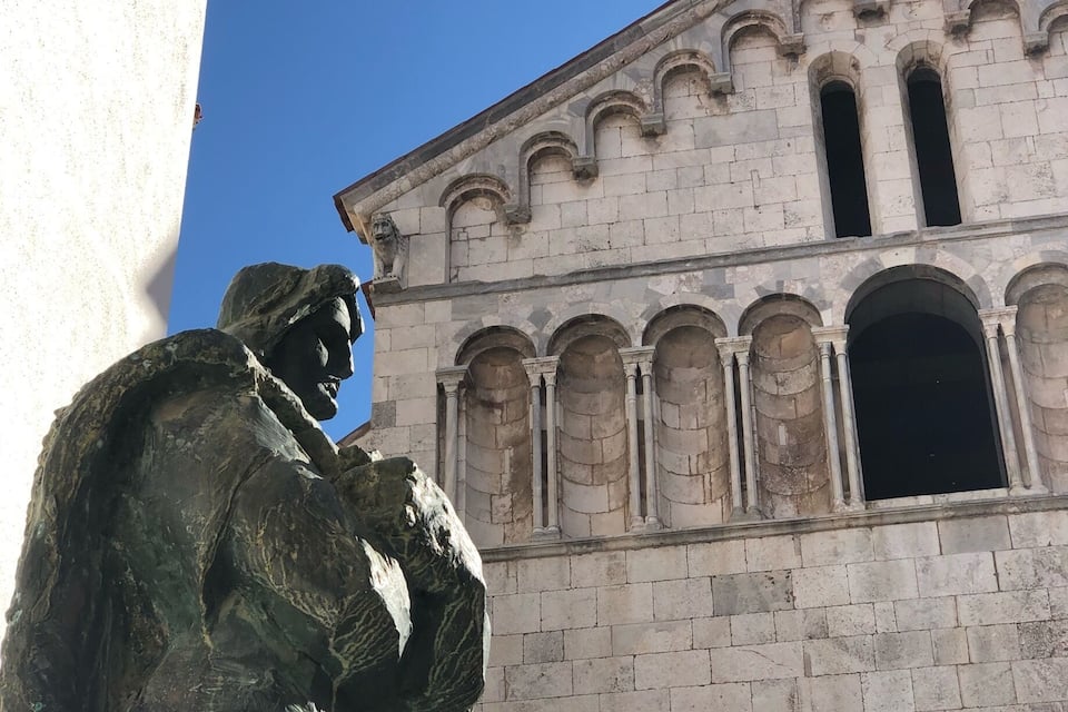 Eine Statue eines Mannes im Anzug und Krawatte, ein Gebäude mit Rundbogenfenstern und ein klarer, blauer Himmel.