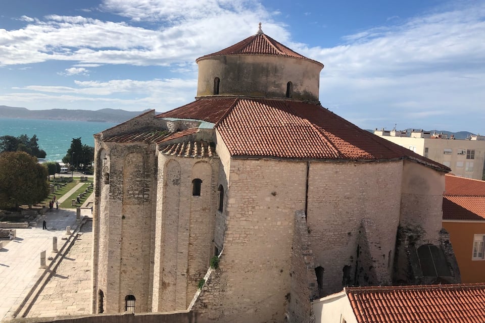Eine historische Kirche mit rotem Ziegeldach und einem Glockenturm, mit Blick auf eine Küstenlandschaft unter blauem Himmel.