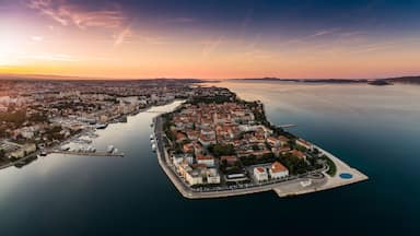 An aerial view of a coastal city with a prominent peninsula, a marina, and a calm sea during sunset.