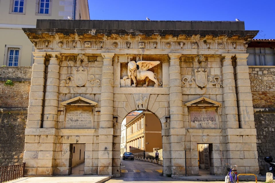 An ornate stone archway with sculptures and inscriptions, leading to a building.