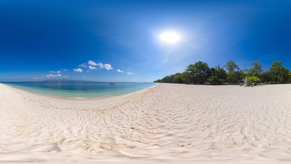 A wide sandy beach with clear turquoise water, a boat in the distance, and a clear blue sky.