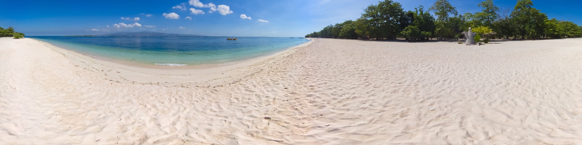 A wide sandy beach with clear turquoise water, a boat in the distance, and a clear blue sky.