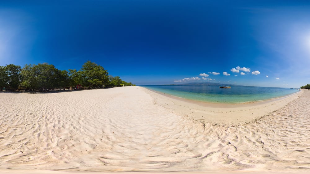 A wide sandy beach with clear turquoise water, a boat in the distance, and lush green trees on the left.