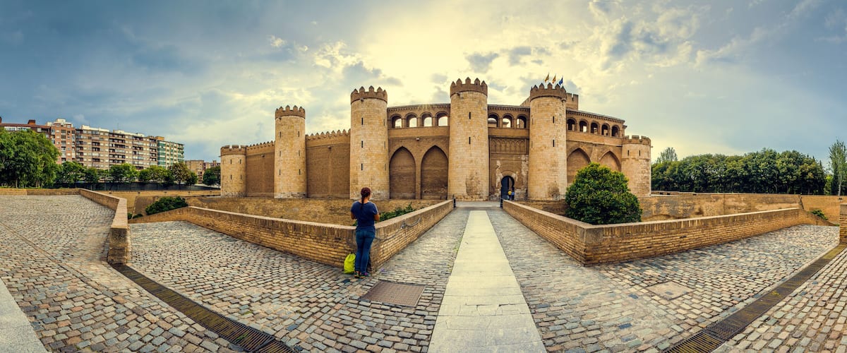 A historic castle with multiple towers and arched windows, surrounded by a cobblestone courtyard and modern buildings in the background.