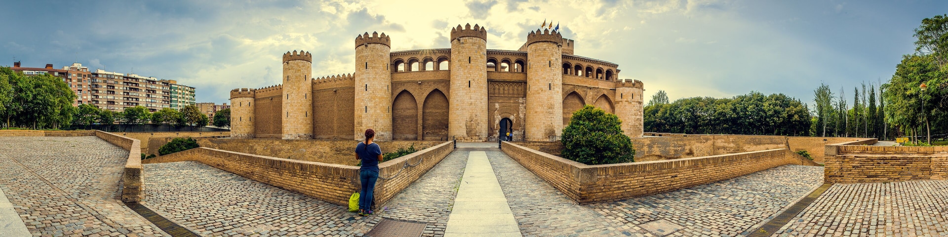 A historic castle with multiple towers and arched windows, surrounded by a cobblestone courtyard and modern buildings in the background.