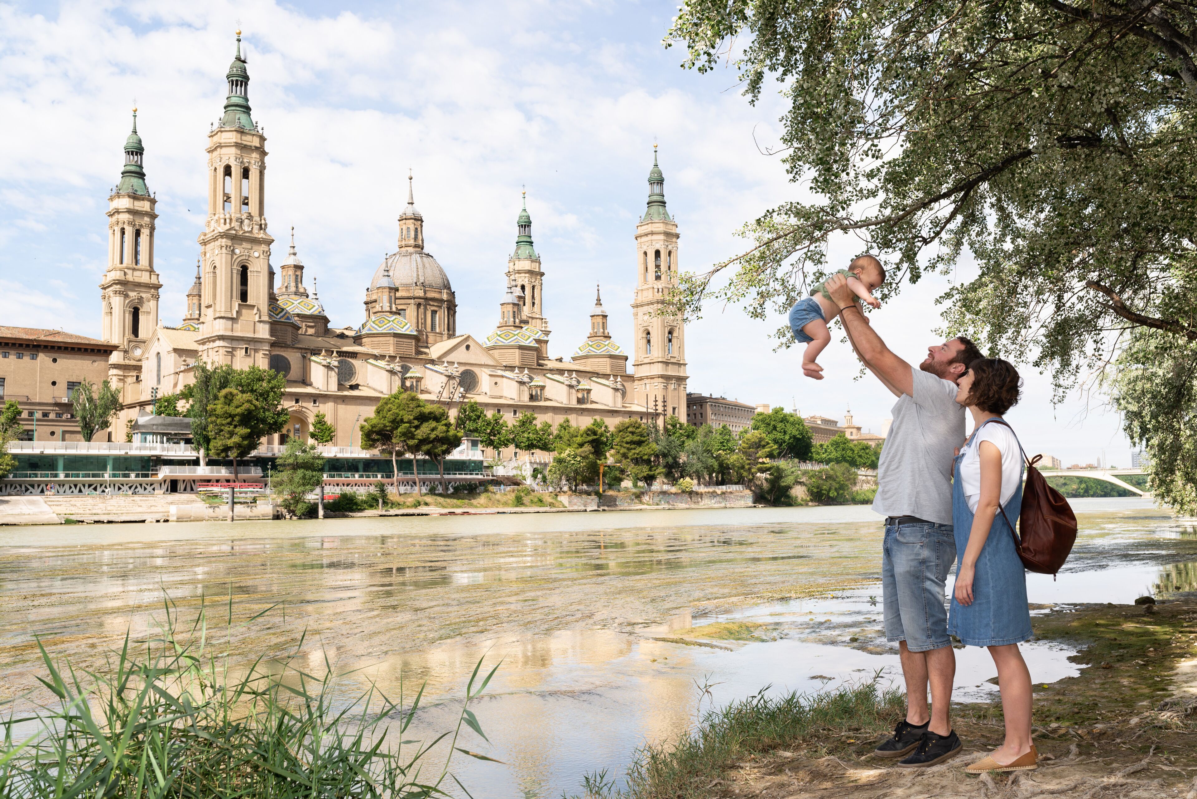 Una catedral histórica con múltiples agujas y cúpulas, un río en primer plano, y una pareja con un niño cerca de la orilla.