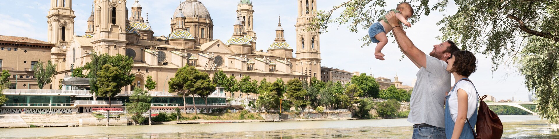 Una catedral histórica con múltiples agujas y cúpulas, un río en primer plano, y una pareja con un niño cerca de la orilla.
