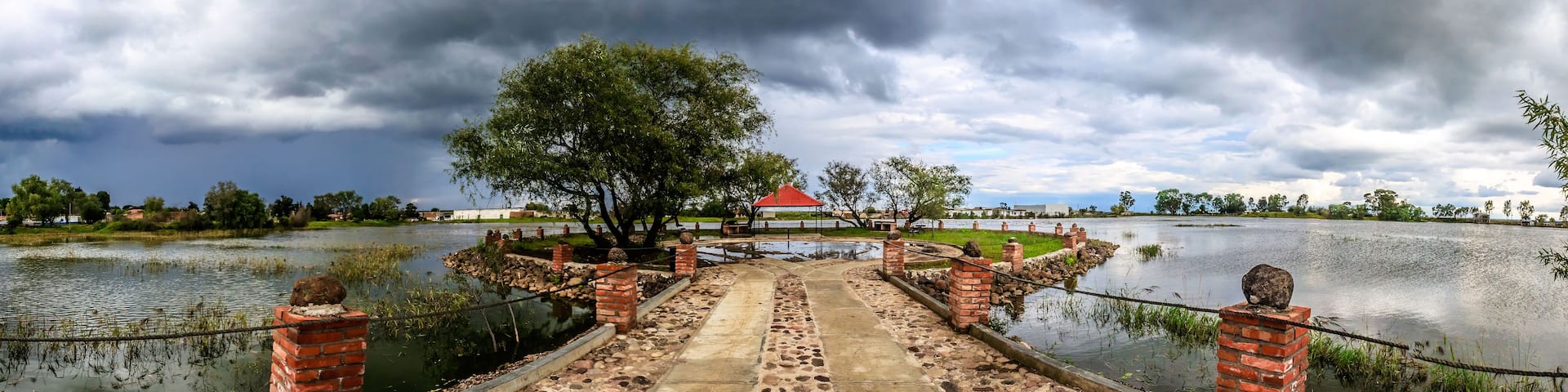 A bridge leading to a park with a red-roofed pavilion, surrounded by water and trees under a cloudy sky.