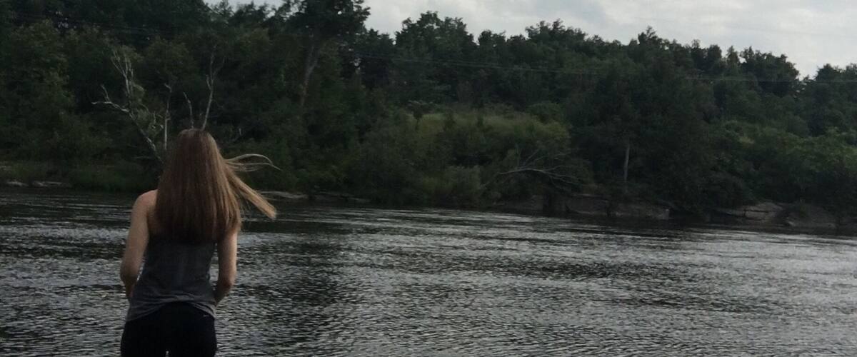 A person walking a dog by a calm river with trees in the background.