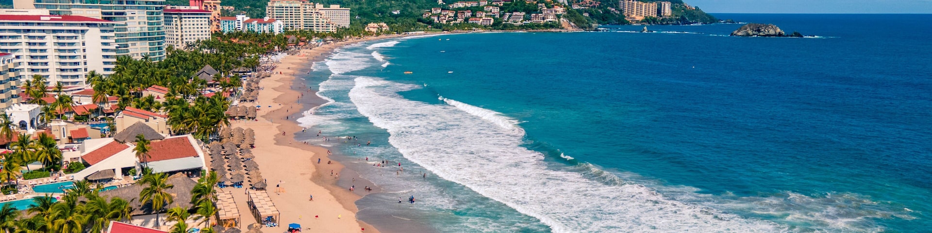 Un paisaje costero con playa, palmeras y montañas al fondo.
