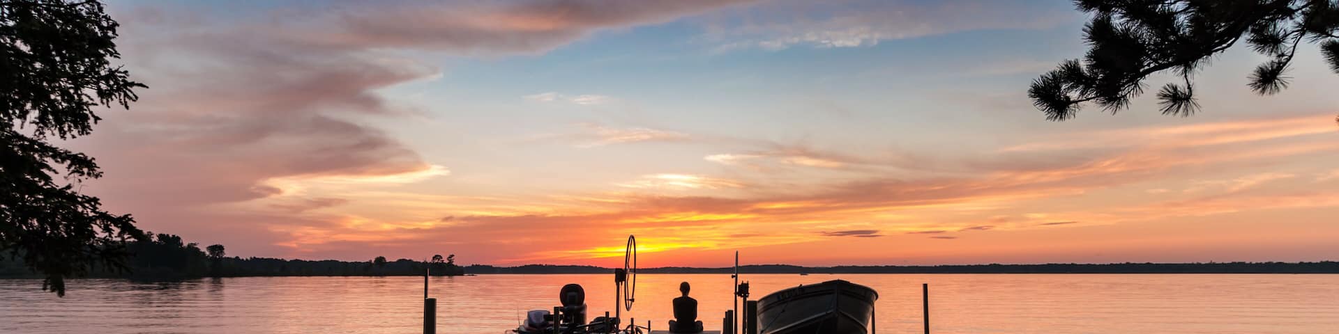 A wooden pier extending into calm water, with a boat docked at the end and a person sitting on the pier.