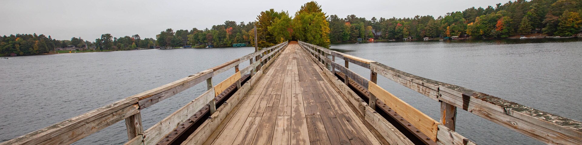 Un muelle de madera se extiende sobre un lago tranquilo, rodeado de árboles con follaje otoñal.